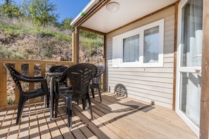 A covered outdoor patio with a table and chairs, wooden deck, and a window with blinds.
