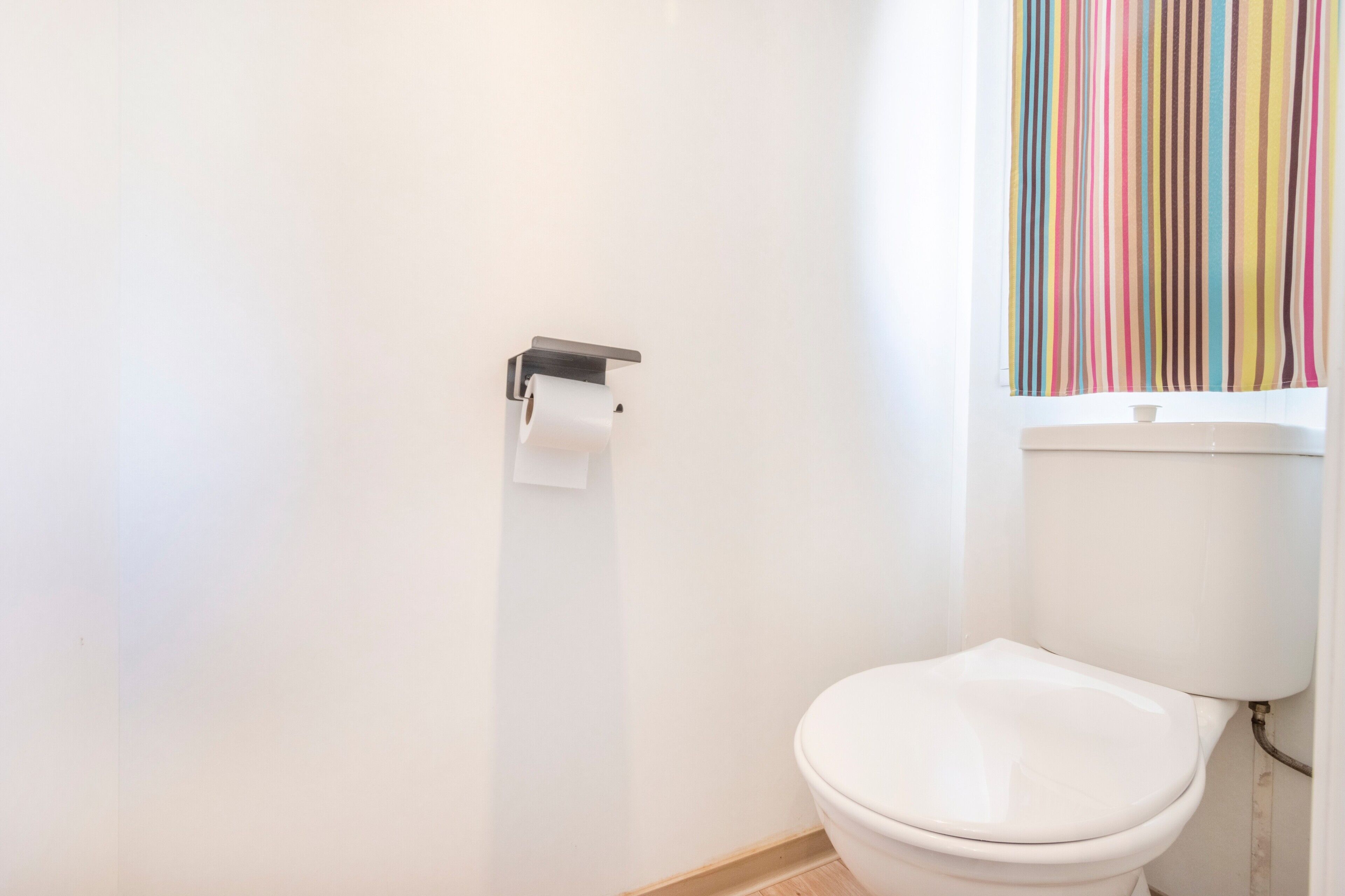 Eine weiße Toilette mit Toilettenpapier auf einem Halter, ein Fenster mit vertikalen Jalousien und ein gefliester Boden.