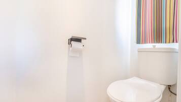 Eine weiße Toilette mit Toilettenpapier auf einem Halter, ein Fenster mit vertikalen Jalousien und ein gefliester Boden.