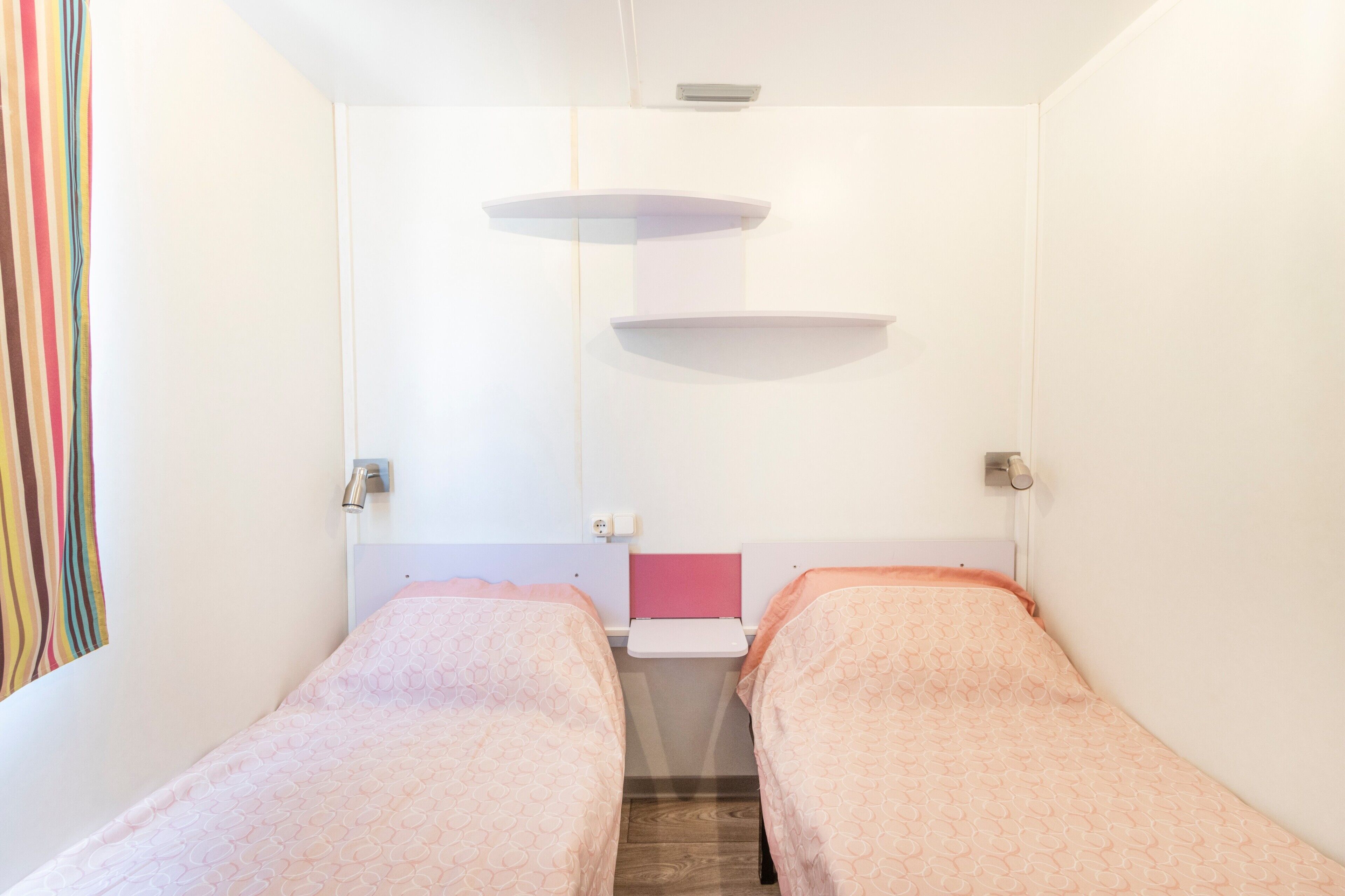 A small, brightly lit room with two beds, a small table, and a wall-mounted shelf.