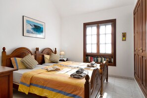 Une chambre à coucher avec un lit, une tête de lit en bois, une fenêtre avec des rideaux, une table de chevet avec une lampe et une bibliothèque.