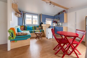 Een knus woongedeelte met kitchenette, een bank met kleurrijke kussens, een kleine eettafel met rode stoelen en een raam met gordijnen.