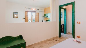 A room with a bed, a green dresser, a mirror, and a view of a kitchen through an open door.