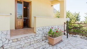 A building entrance with a stone-paved porch, a wooden door, and a potted plant with flowers.