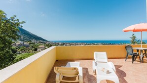 A balcony with white plastic chairs and a table, a red umbrella, and a view of the sea and distant buildings. - Apartment 'Appartamento Superior Con Terrazza5' with Shared Pool, Wi-Fi and Air Conditioning (Castellammare del Golfo)