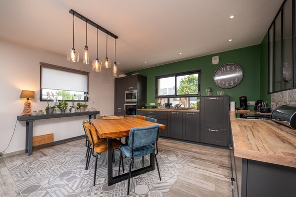 Une cuisine moderne avec un coin repas, comprenant une table en bois, des chaises, des luminaires suspendus et une grande horloge au mur.