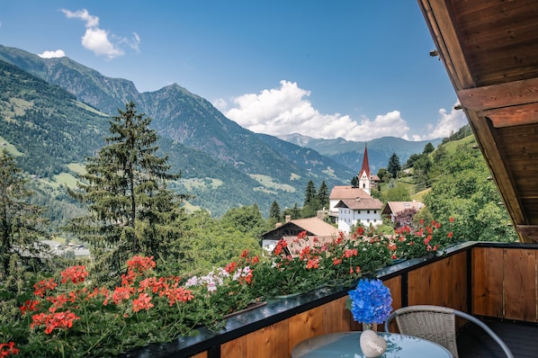 Un balcon en bois avec une table et une chaise, donnant sur un paysage montagneux avec une église et des maisons.
