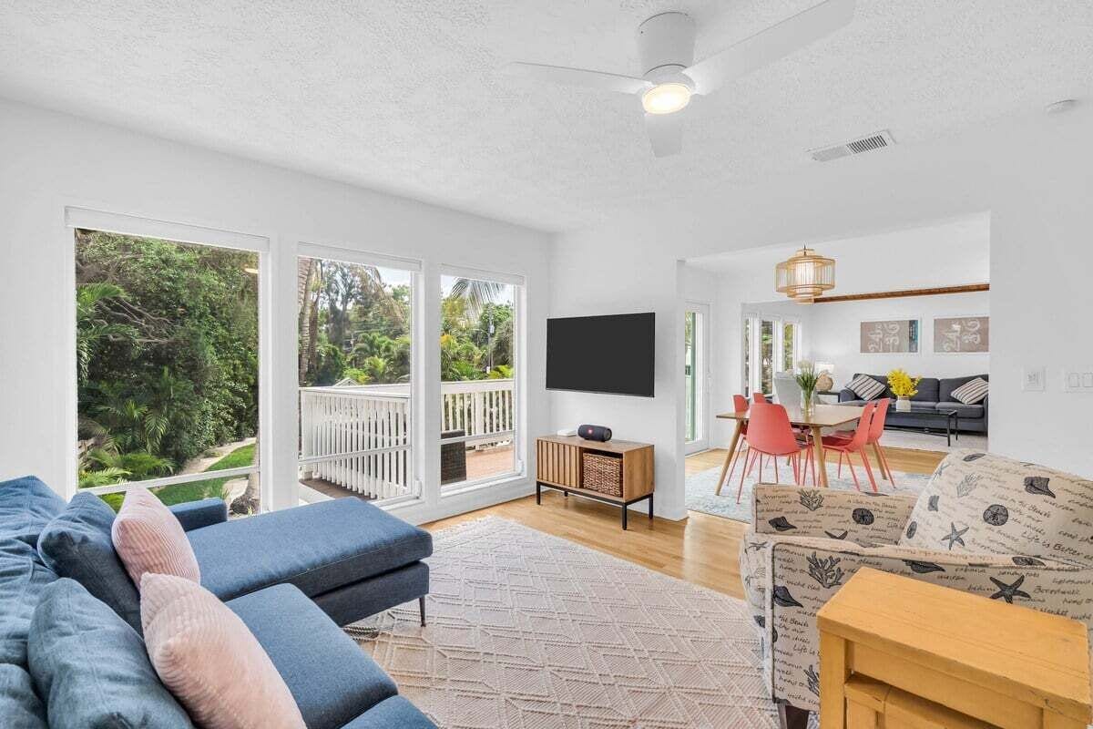 A spacious living area with a large sliding glass door to a balcony, a flat-screen TV mounted on the wall, and a dining area with red chairs.
