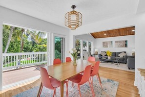 A spacious room with a wooden dining table, red chairs, a sofa, and a balcony with a view of greenery.