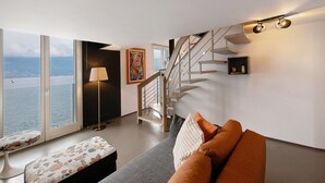 Un salon moderne comprenant un canapé, un banc, une lampe, un escalier et une grande fenêtre donnant sur l’eau.