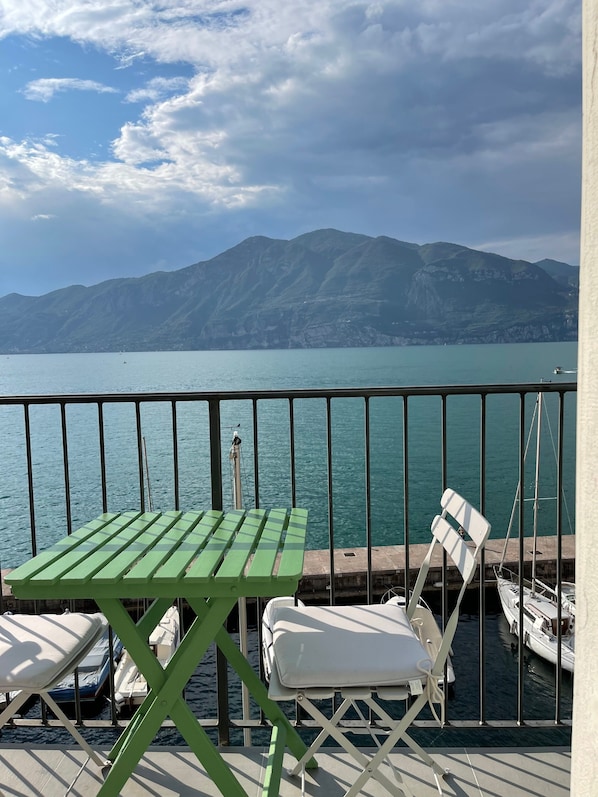 A balcony with a green table and white lounge chairs, overlooking a calm lake with boats and mountains in the background.