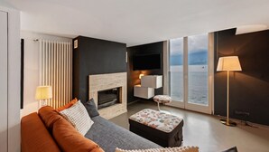 Un salon moderne comprenant un canapé, un pouf, une cheminée, un téléviseur et une vue sur la mer grâce à de grandes fenêtres.