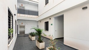 Corredor interno limpo e moderno, com piso ladrilhado, plantas em vasos e um elevador visível.