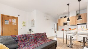 Apartamento moderno com sofá cinza, manta colorida, armários de madeira e área de cozinha.