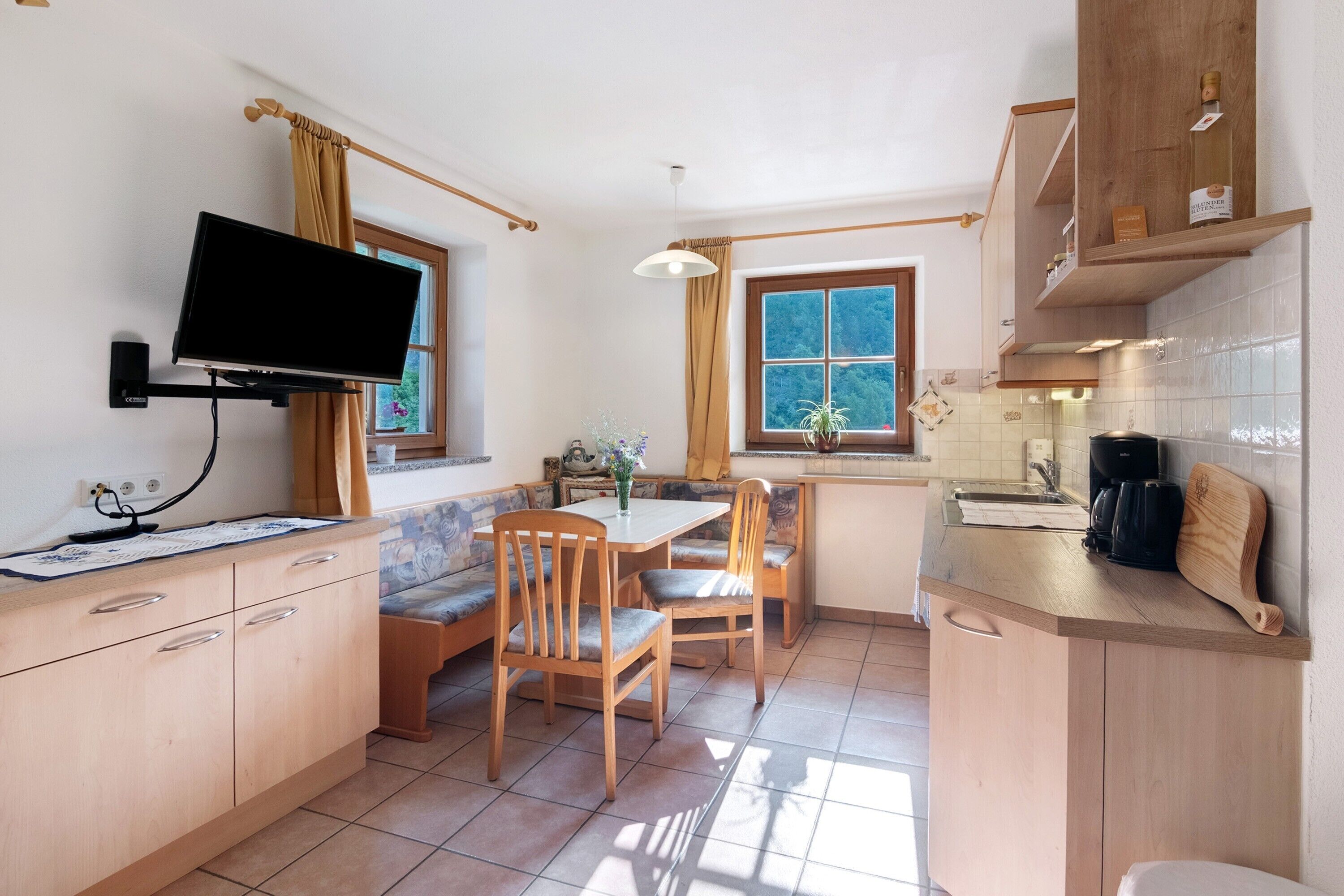 A kitchen with a dining area, wooden cabinets, a TV mounted on the wall, and a window with curtains.