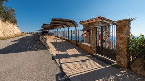 A coastal road with a stone guardrail, a covered walkway, and a stone building with a metal gate.
