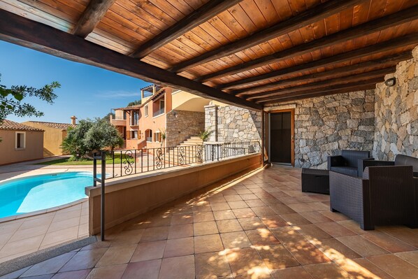 A covered outdoor area with a swimming pool, stone wall, and outdoor furniture.