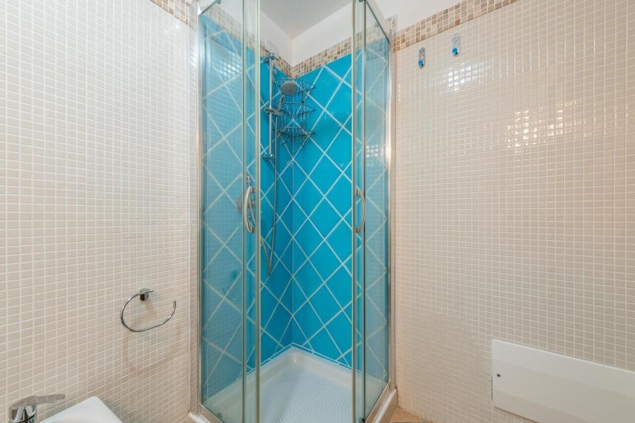 A glass-enclosed shower with a blue-tiled wall, a white sink, and a beige tiled floor.