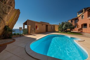 Una piscina con bordo curvo, una grande formazione rocciosa e un edificio con tetto in tegole e balconi.