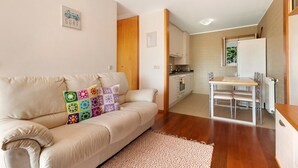 Una sala de estar moderna con un sofá beige, un cojín colorido, una mesa de comedor con sillas, una zona de cocina y un ventanal pequeño.