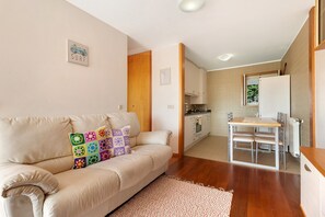 Una sala de estar moderna con un sofá beige, un cojín colorido, una mesa de comedor con sillas, una zona de cocina y un ventanal pequeño.