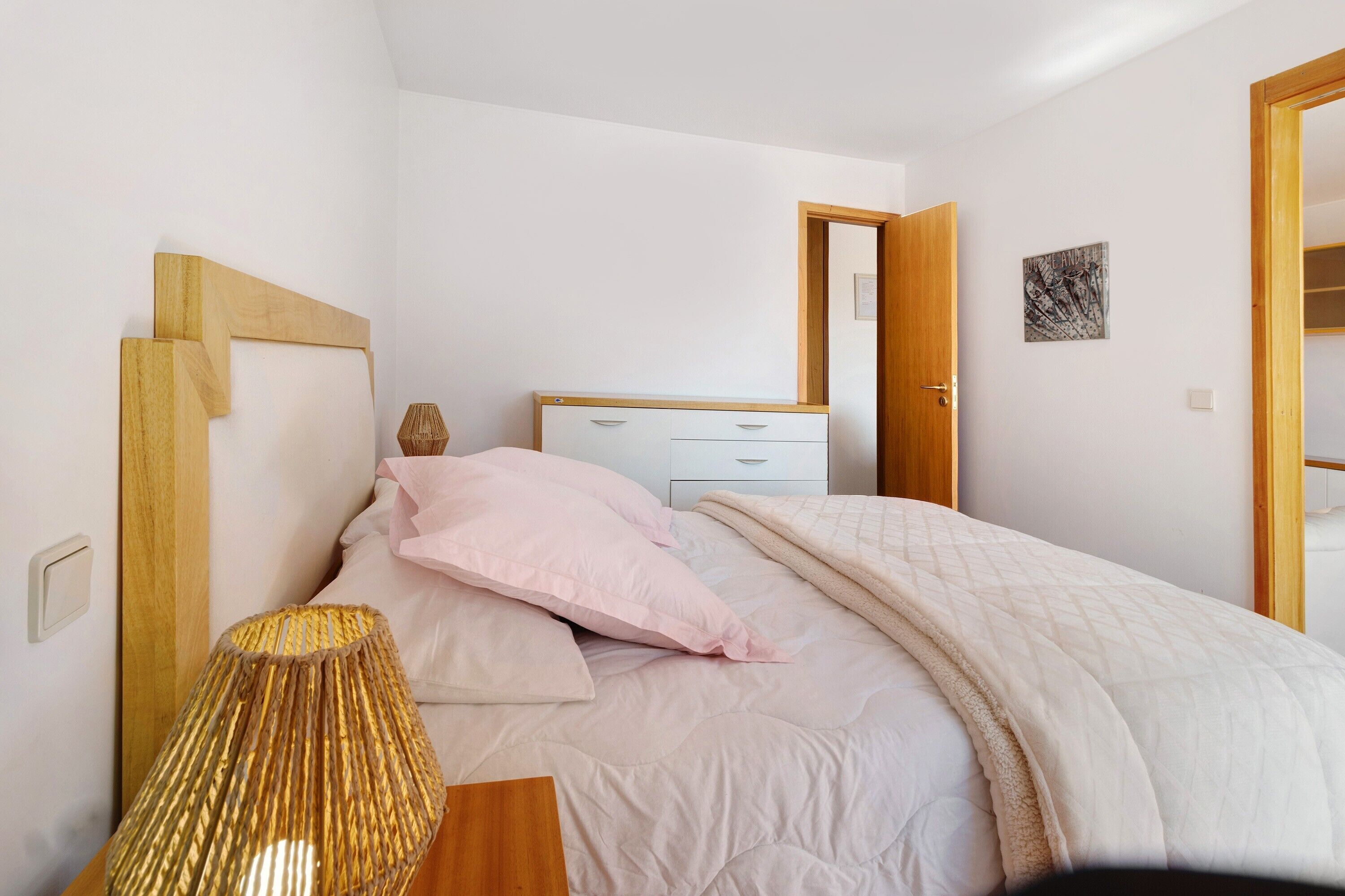 A bedroom with a bed, bedside table, lamp, dresser, and a door leading to another room.