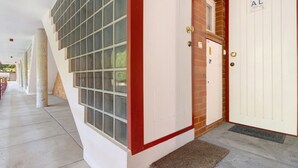 Entrada de un edificio con una puerta blanca marcada 'AL', una ventana con marco rojo y piso de concreto.