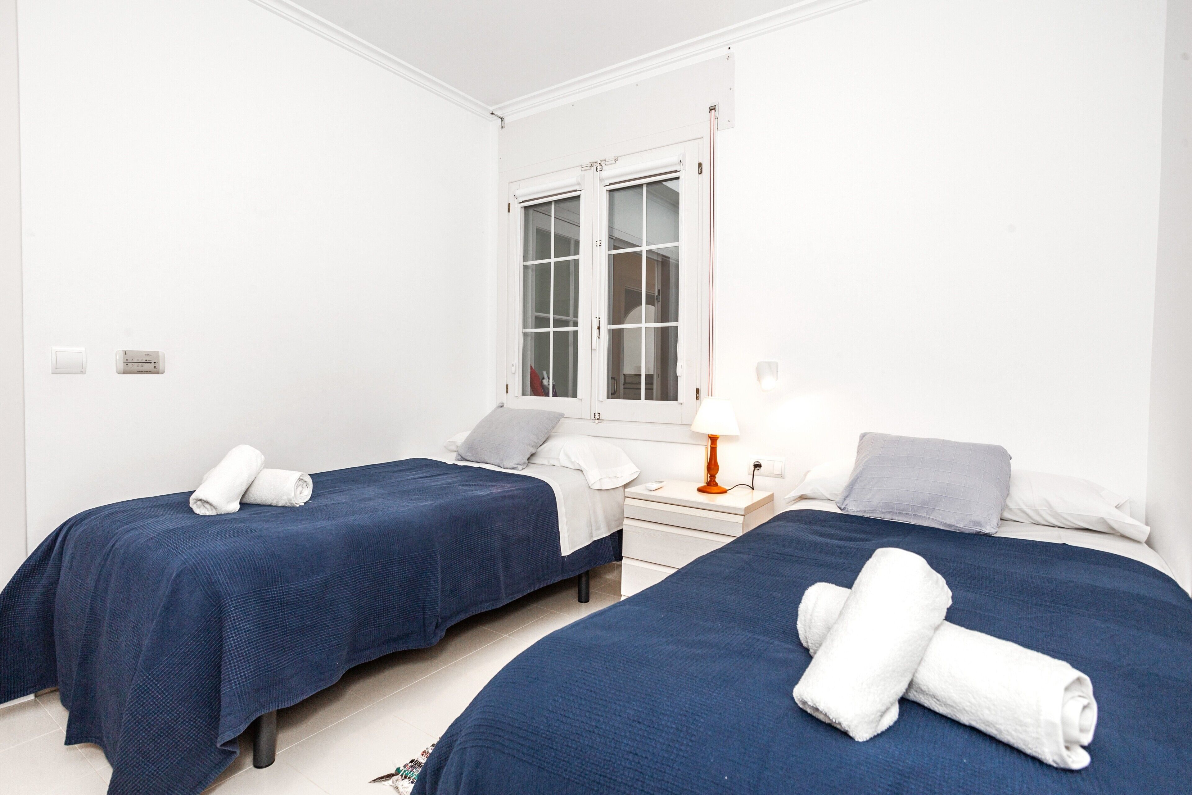 Une chambre avec deux lits, chacun recouvert d’une couvre-lit bleue et agrémenté d’oreillers blancs, une table de chevet avec une lampe, et une fenêtre donnant sur un paysage agréable.