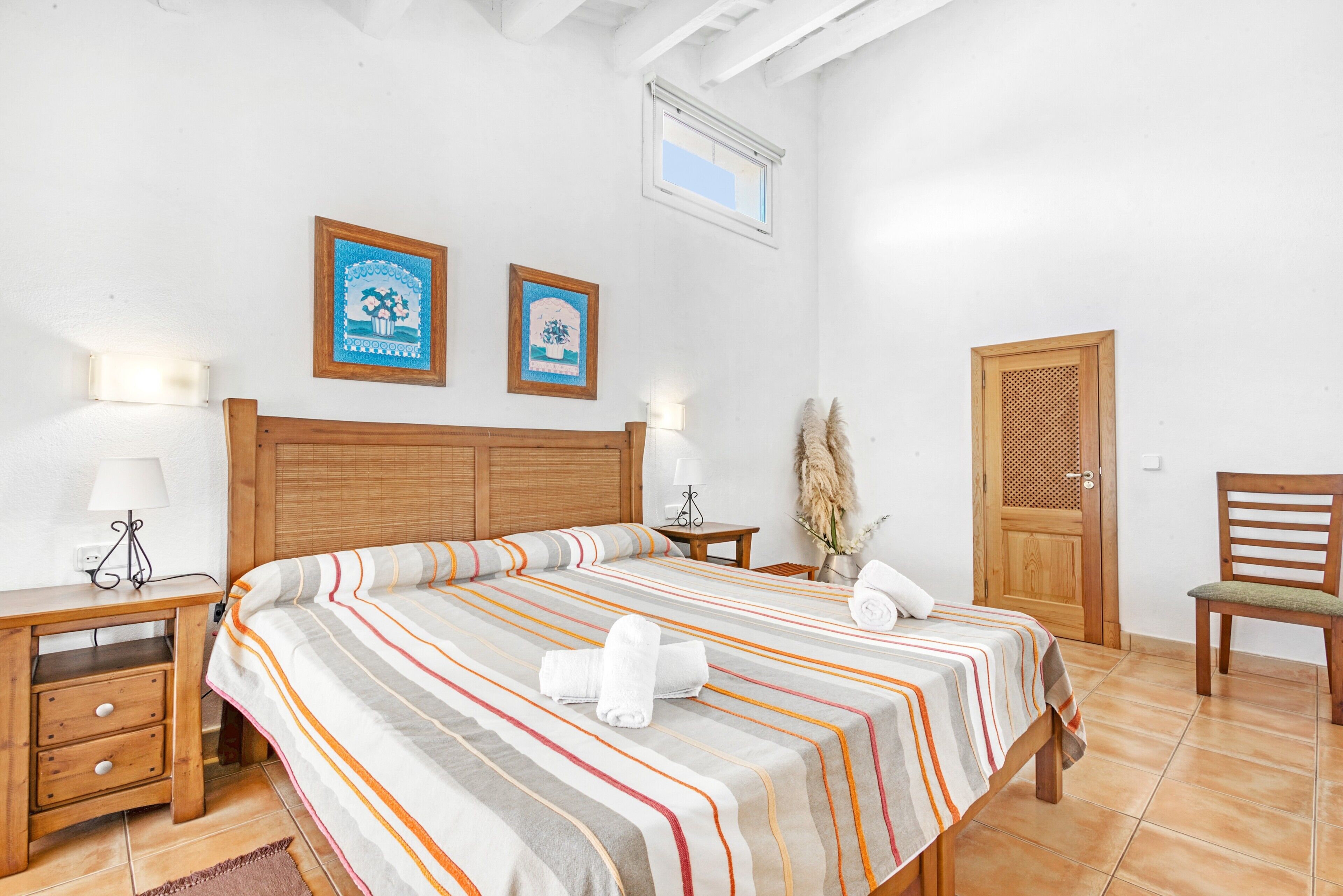 Un dormitorio con una cama grande, cabecera de madera, dos mesitas de noche con lámparas, una puerta de madera y una silla.