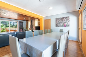 Un espace de repas moderne avec une longue table et des chaises, une cuisine en arrière-plan et une grande fenêtre donnant sur un paysage verdoyant.