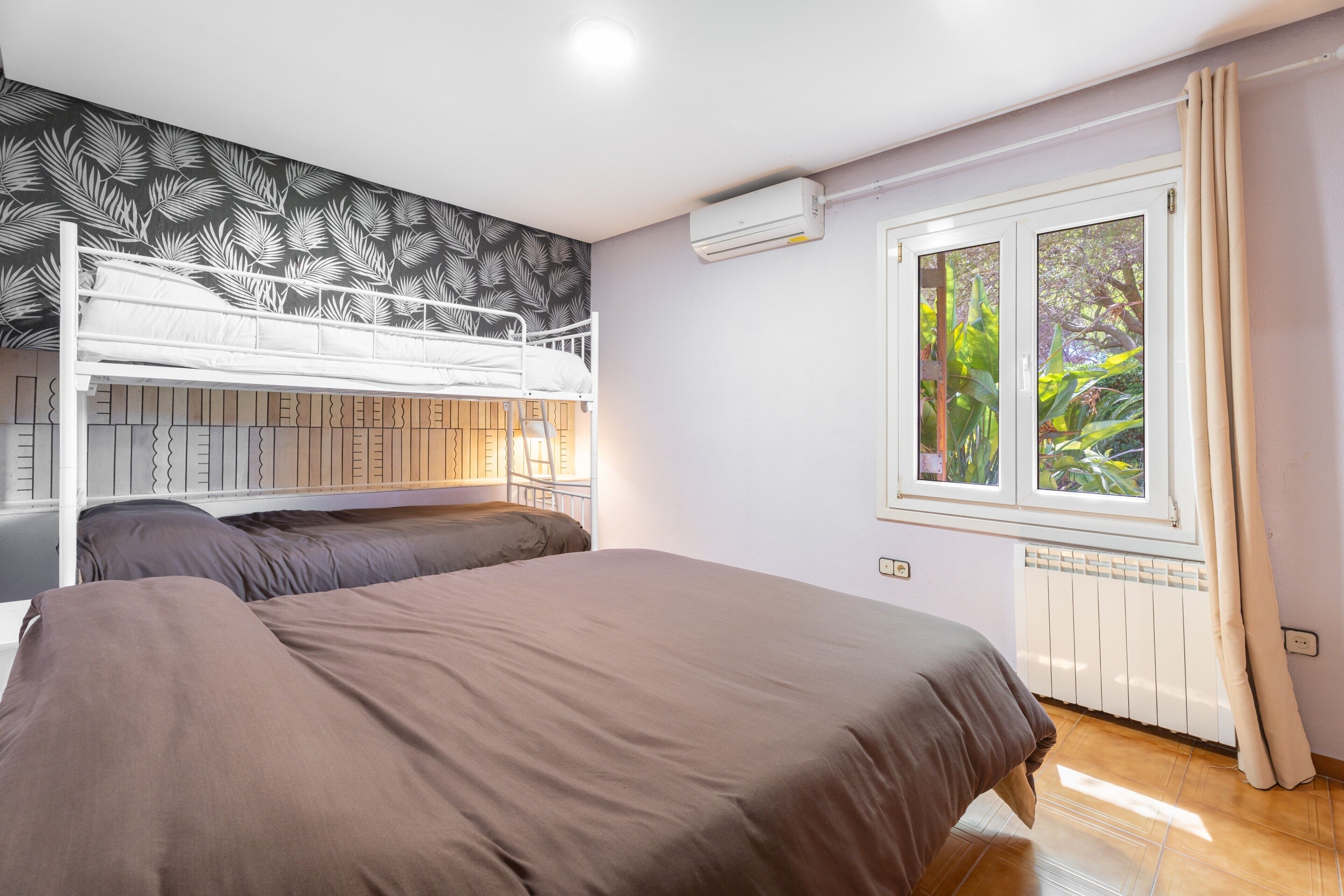 Une chambre avec des lits superposés, une fenêtre donnant sur un espace verdoyant, un appareil de climatisation et un radiateur.