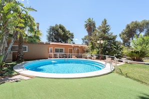 Une piscine entourée d’une terrasse, un bâtiment à la façade marron et divers arbres.