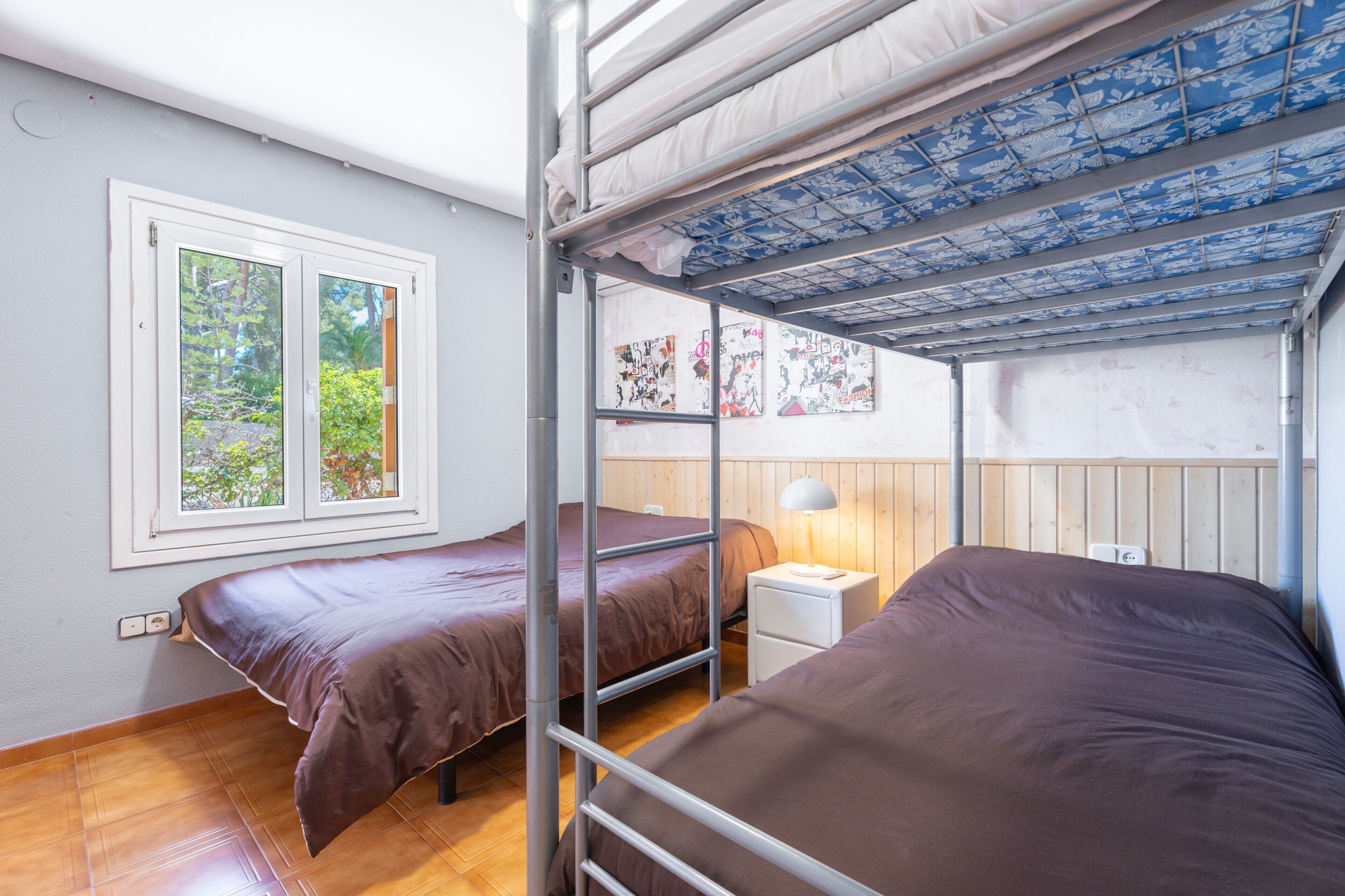 Een studentenkamer met stapelbedden, een raam met uitzicht op groen, een nachtkastje met lamp, en een muur met kunstwerken.