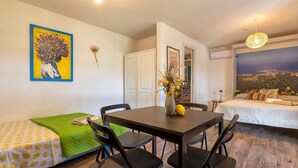 A room with a bed, a dining table with chairs, a painting on the wall, and a view of a landscape through a window.