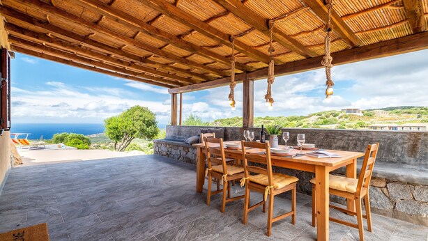 An outdoor wooden table with chairs, a stone wall, and a view of the sea.