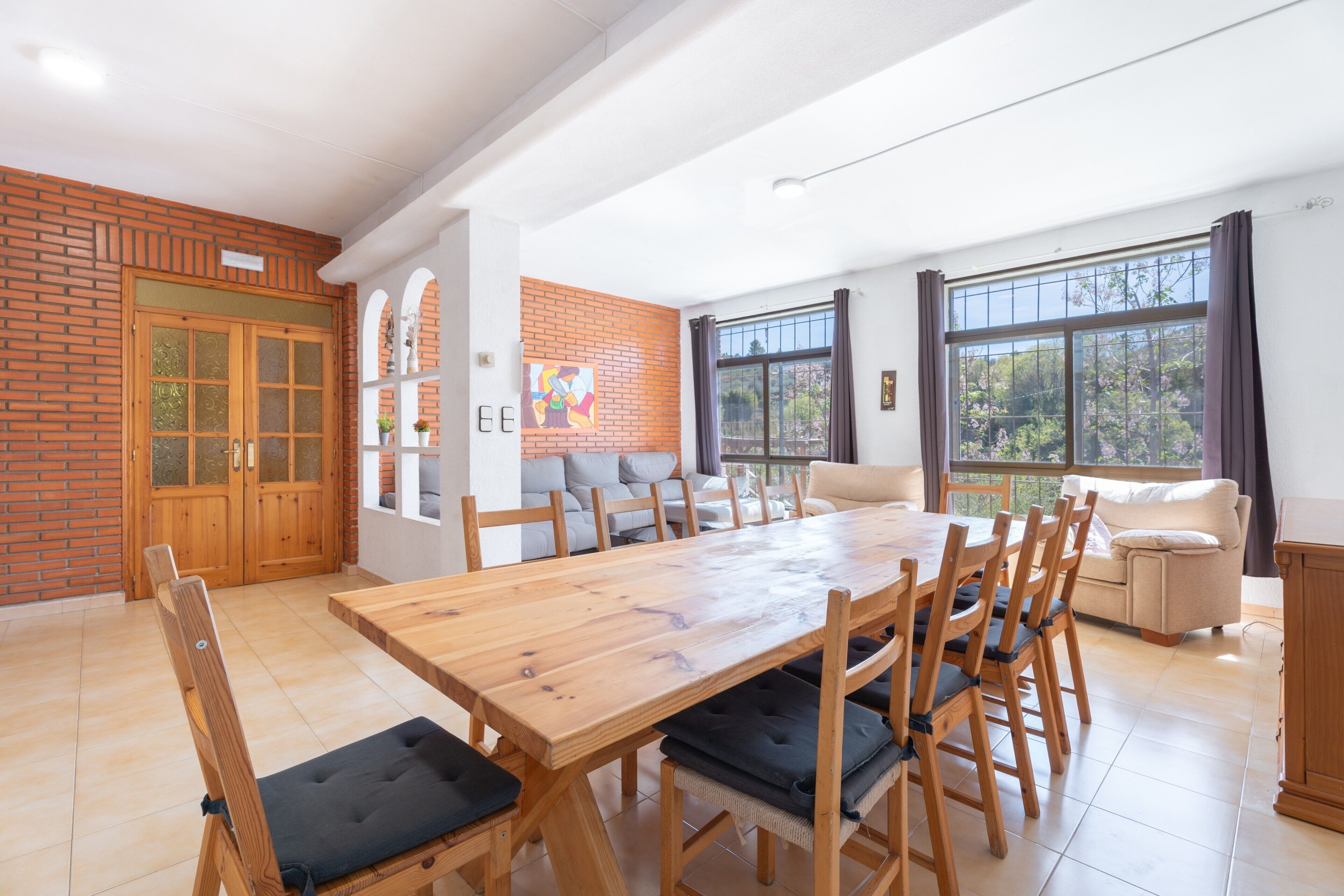 A dining area with a wooden table and chairs, a brick wall, a sofa, and large windows.
