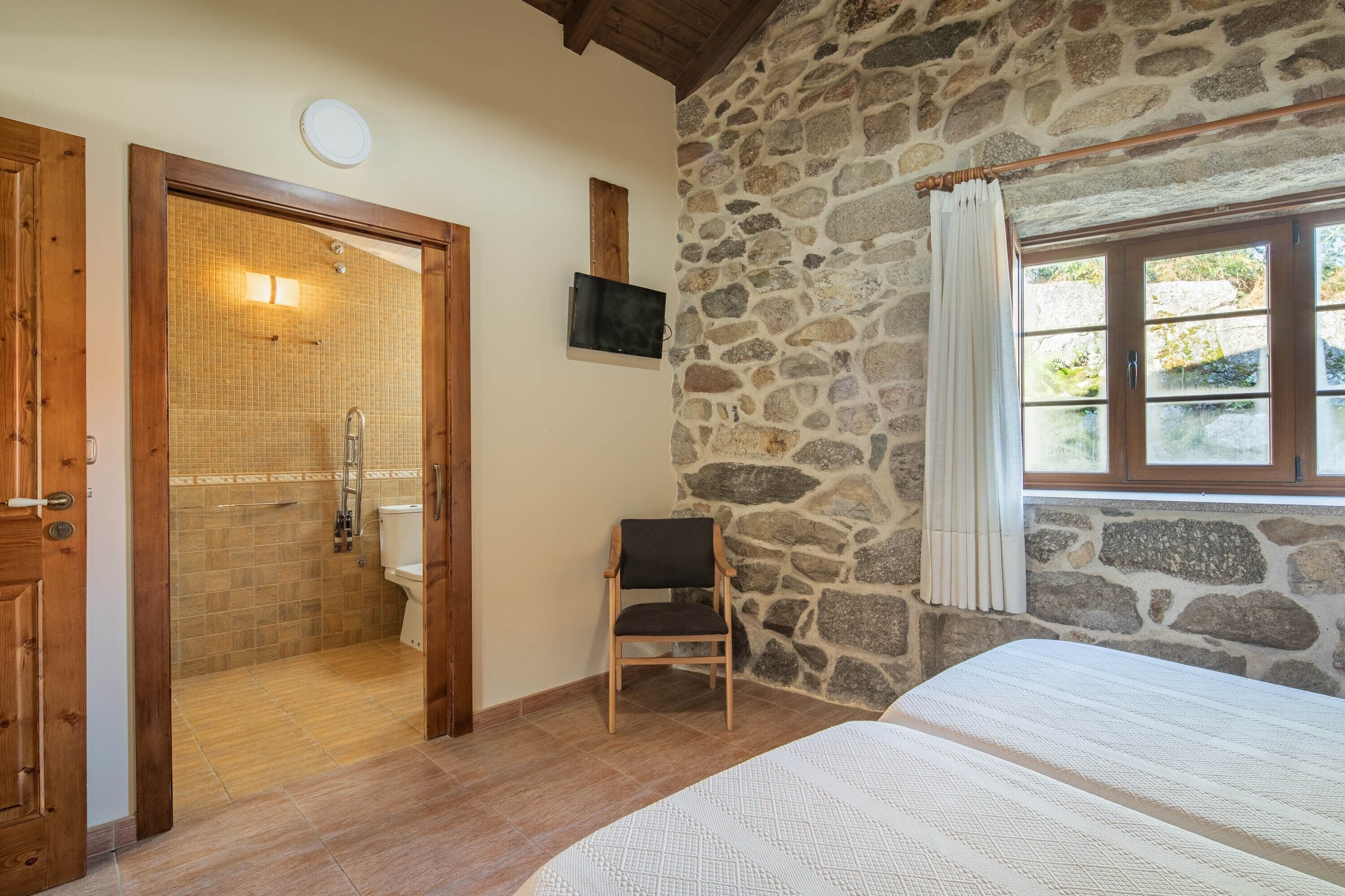 Una habitación con pared de piedra, puerta de madera, ducha, cama y una silla.