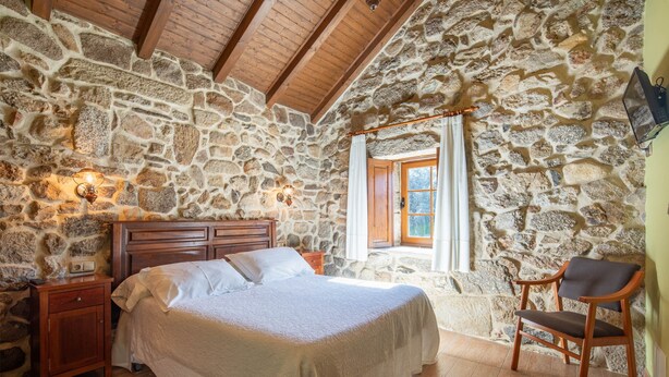 Une chambre avec un mur en pierre, des poutres en bois au plafond, un lit, une table de chevet, une chaise et une fenêtre avec des rideaux.