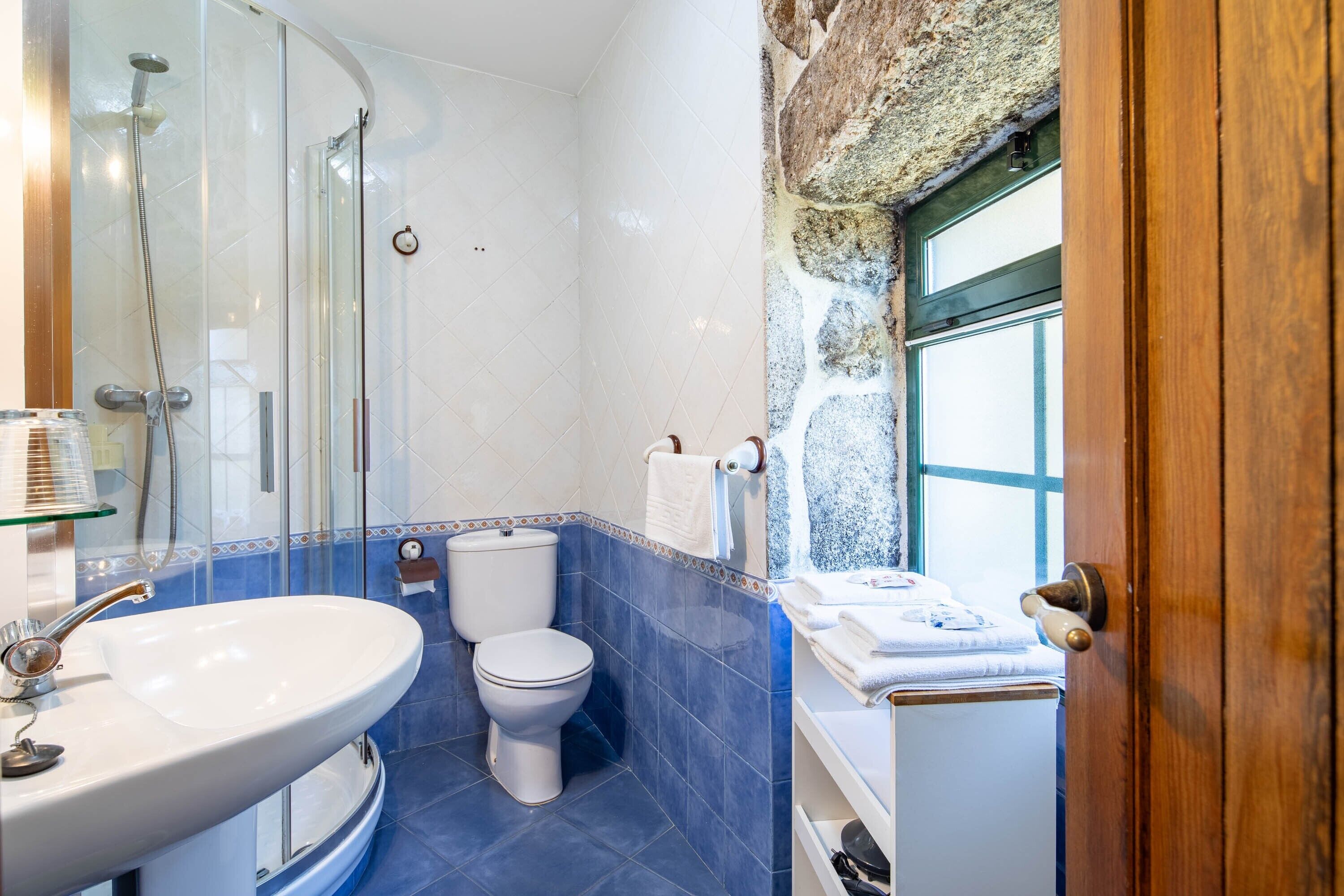 A bathroom with a glass shower enclosure, a white sink, a toilet, and a window with a stone wall.