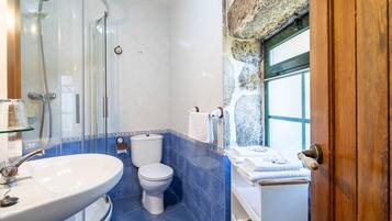 A bathroom with a glass shower enclosure, a white sink, a toilet, and a window with a stone wall.