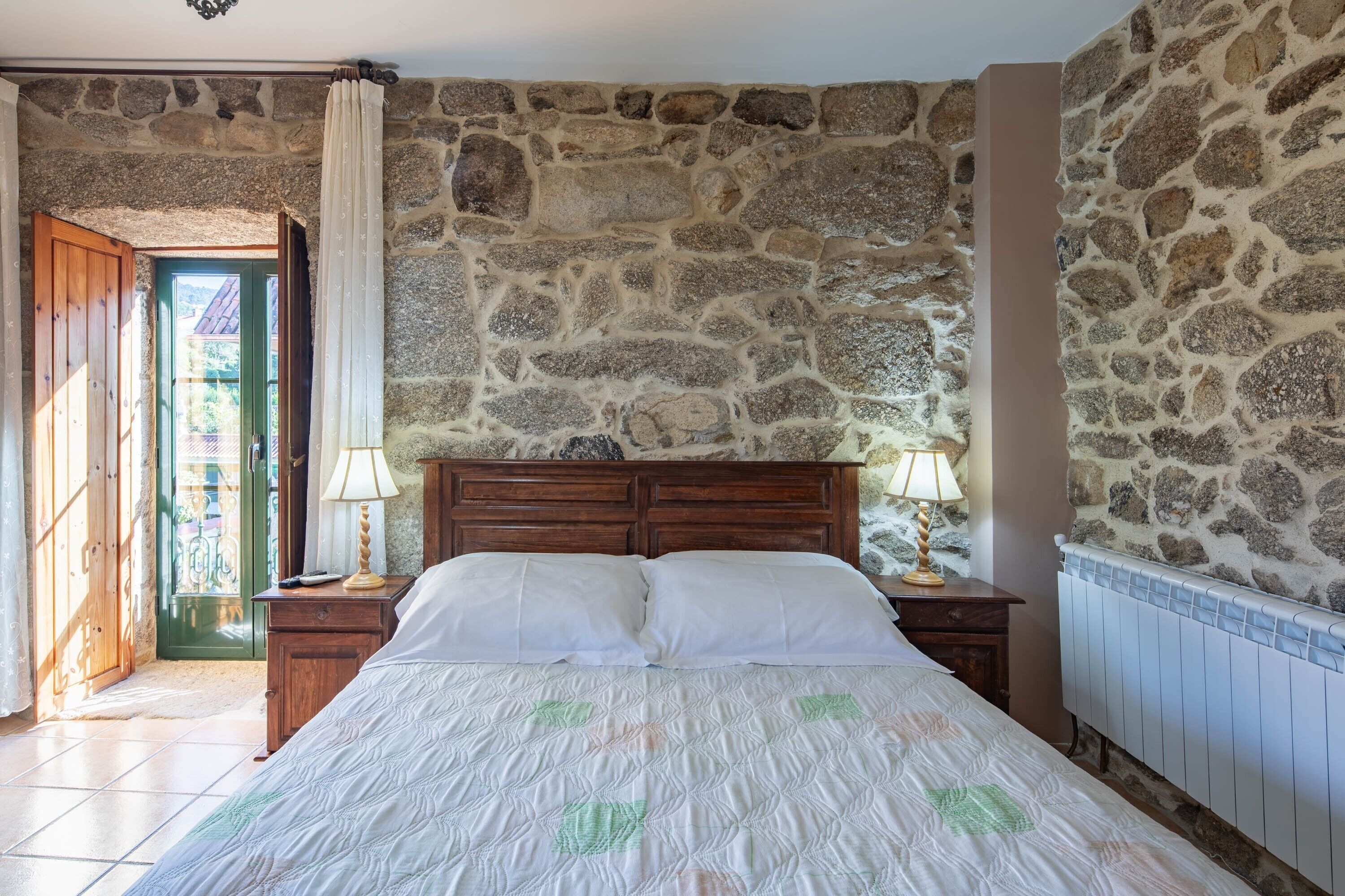 A bedroom with a stone wall, wooden bed frame, two bedside lamps, and a radiator.