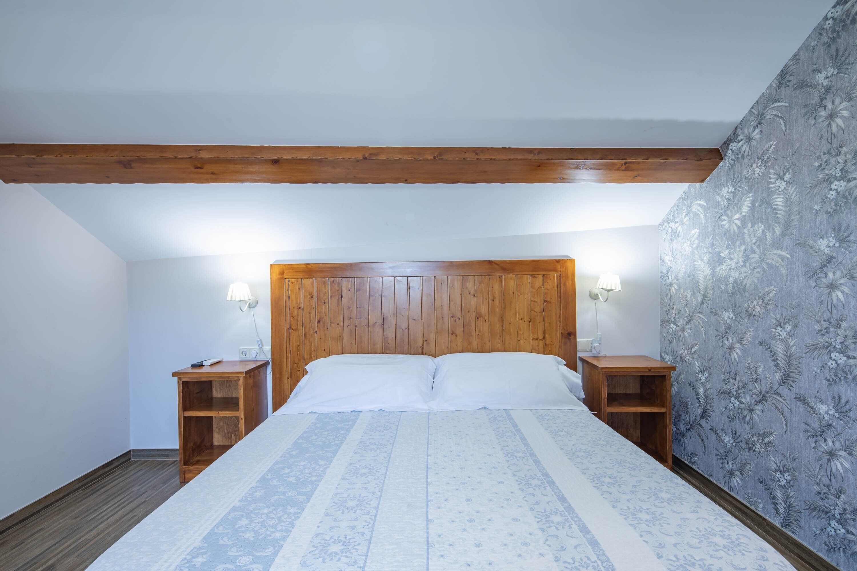 A bedroom with a wooden headboard, two bedside tables with lamps, a patterned bedspread, and floral wallpaper on one wall.