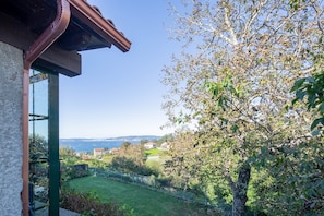 A view from a window with a metal railing, a tree, and a distant landscape including houses and a body of water.