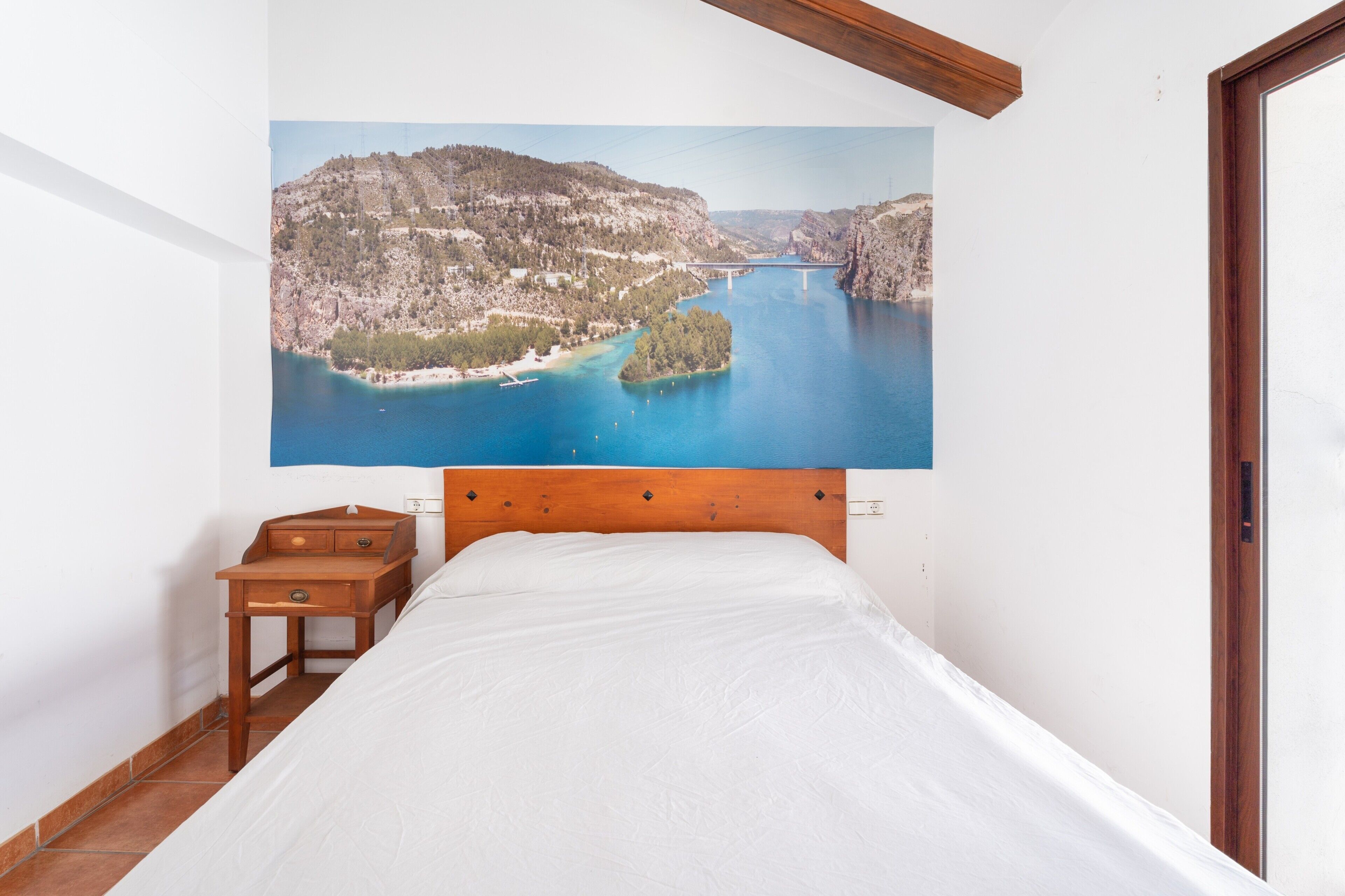 Un dormitorio con cama, mesita de noche de madera, un cuadro de un paisaje costero y una puerta.