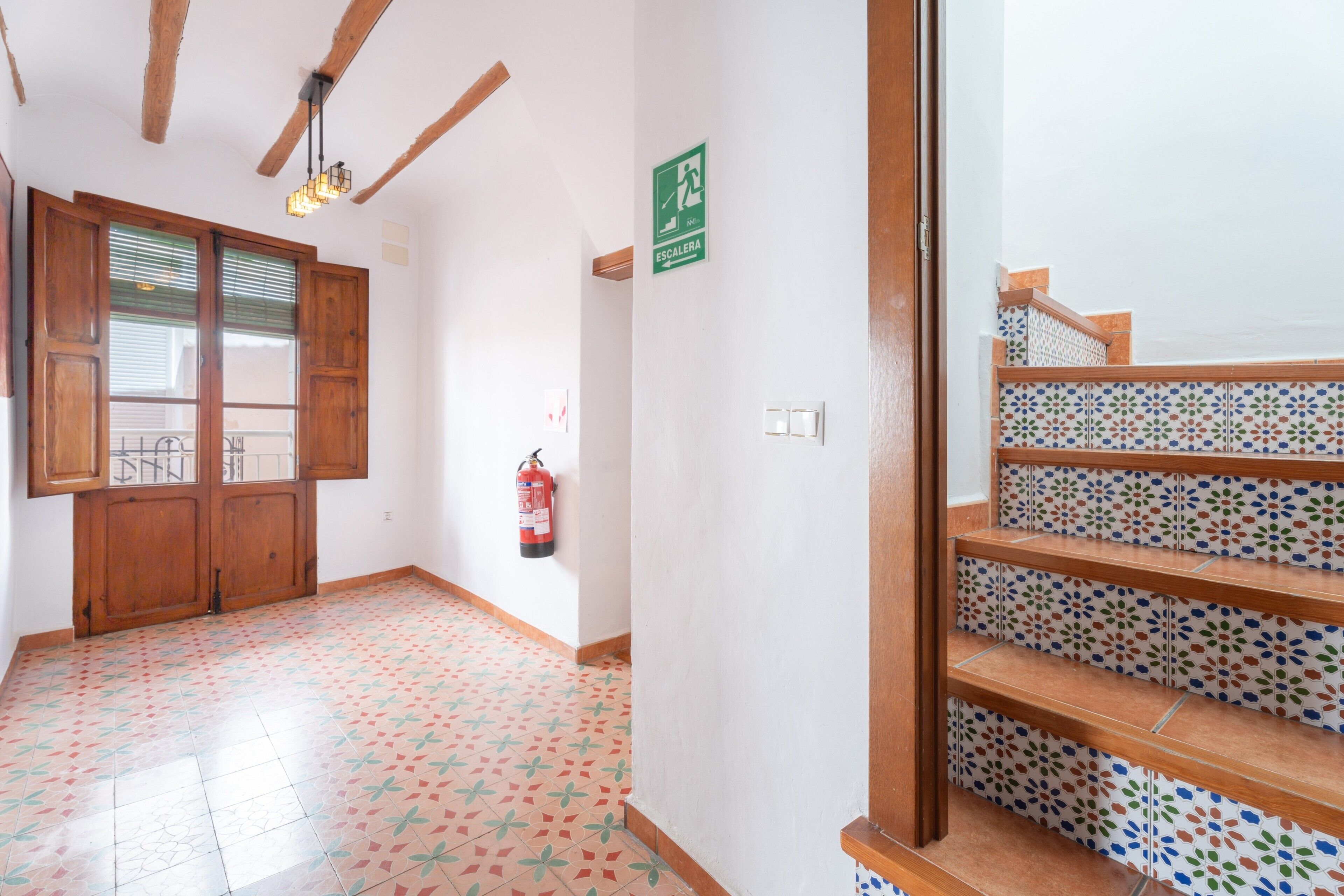 Un pasillo con una puerta de madera, un extintor, un letrero de salida de emergencia verde y una escalera con baldosas estampadas.