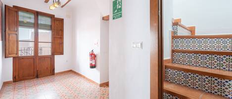 Un pasillo con una puerta de madera, un extintor, un letrero de salida de emergencia verde y una escalera con baldosas estampadas.