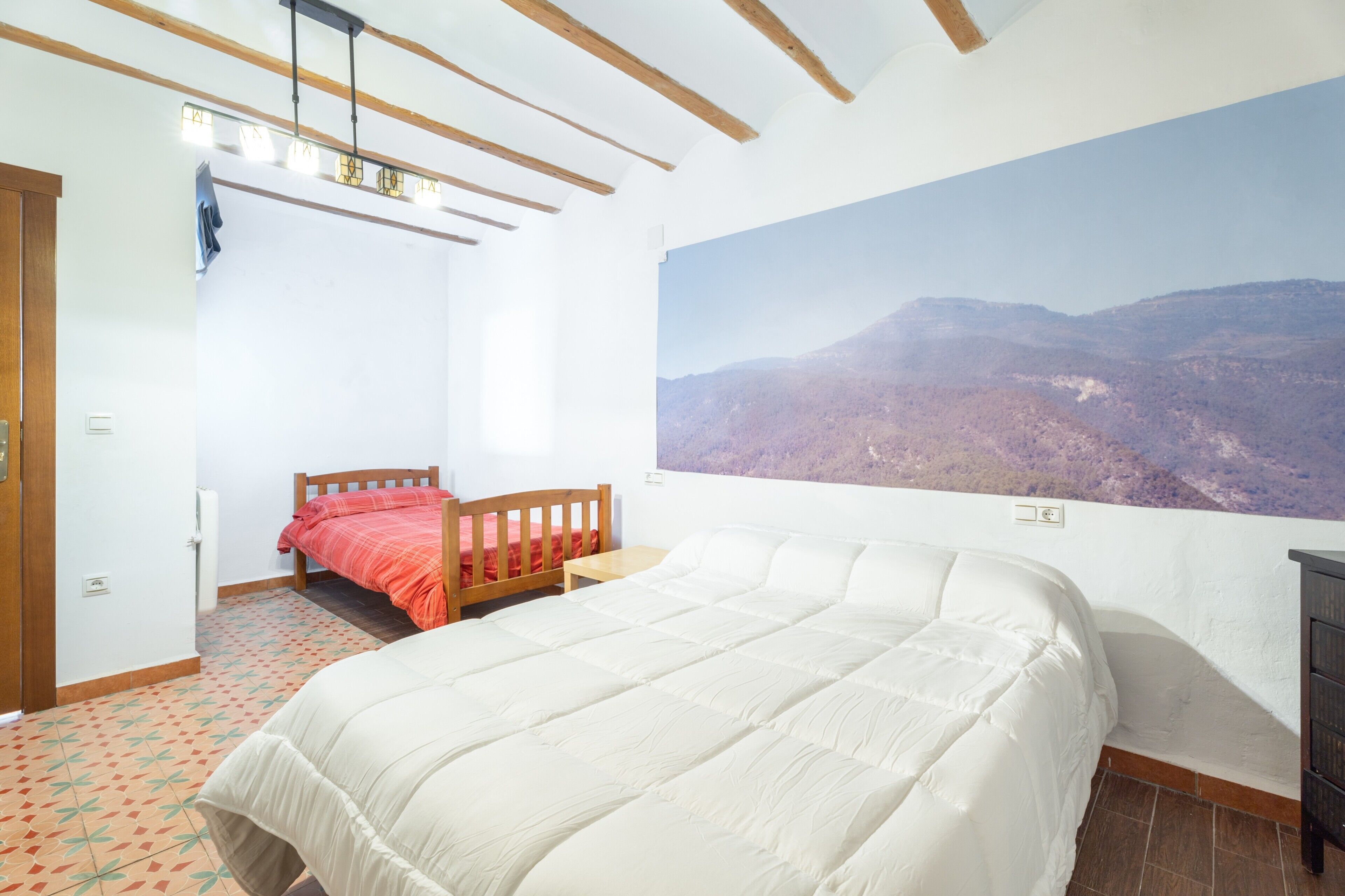 Un dormitorio con una cama grande, una cama más pequeña, cabecera de madera, mesita de noche, estante en la pared y un amplio ventanal con vistas a la montaña.