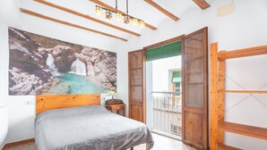 A bedroom with a bed, a wooden headboard, a painting of a waterfall, a small table with a lamp, and a balcony door.