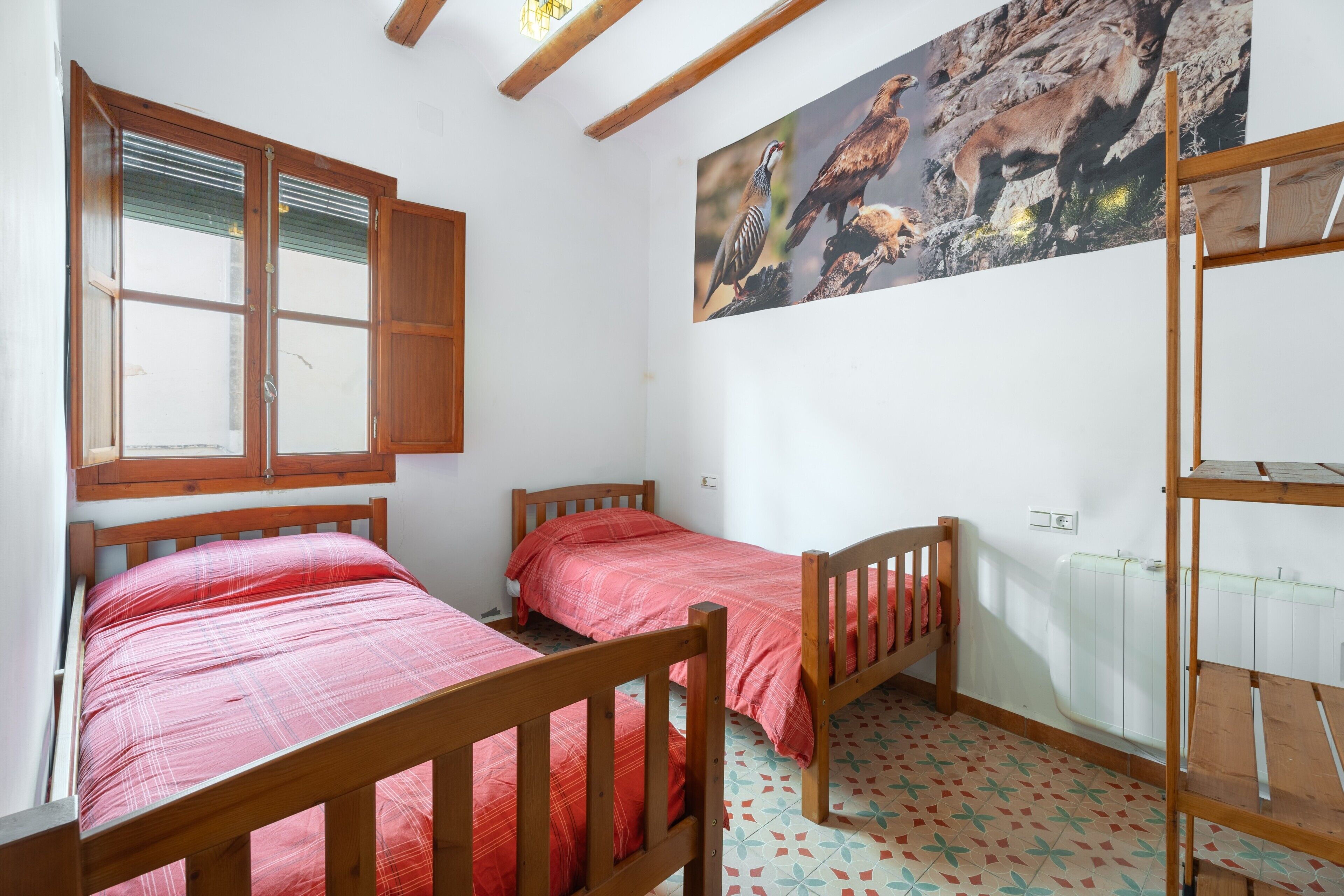 Quarto com duas camas de madeira, janela com persianas, radiador e prateleira fixada na parede.