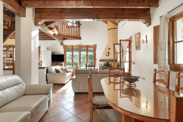 A spacious living area with a glass dining table, wooden chairs, a sofa, and a staircase leading to an upper level.